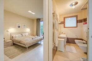 Un dormitorio con cama, mesitas de noche y un velador. Junto al dormitorio, un baño con lavamanos, inodoro y bañera.