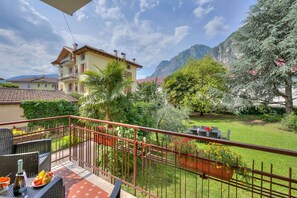 A balcony with wicker furniture, a table with fruit and a bottle, and a view of a mountain and garden. - The Bon Ora Apartment (Riva del Garda)