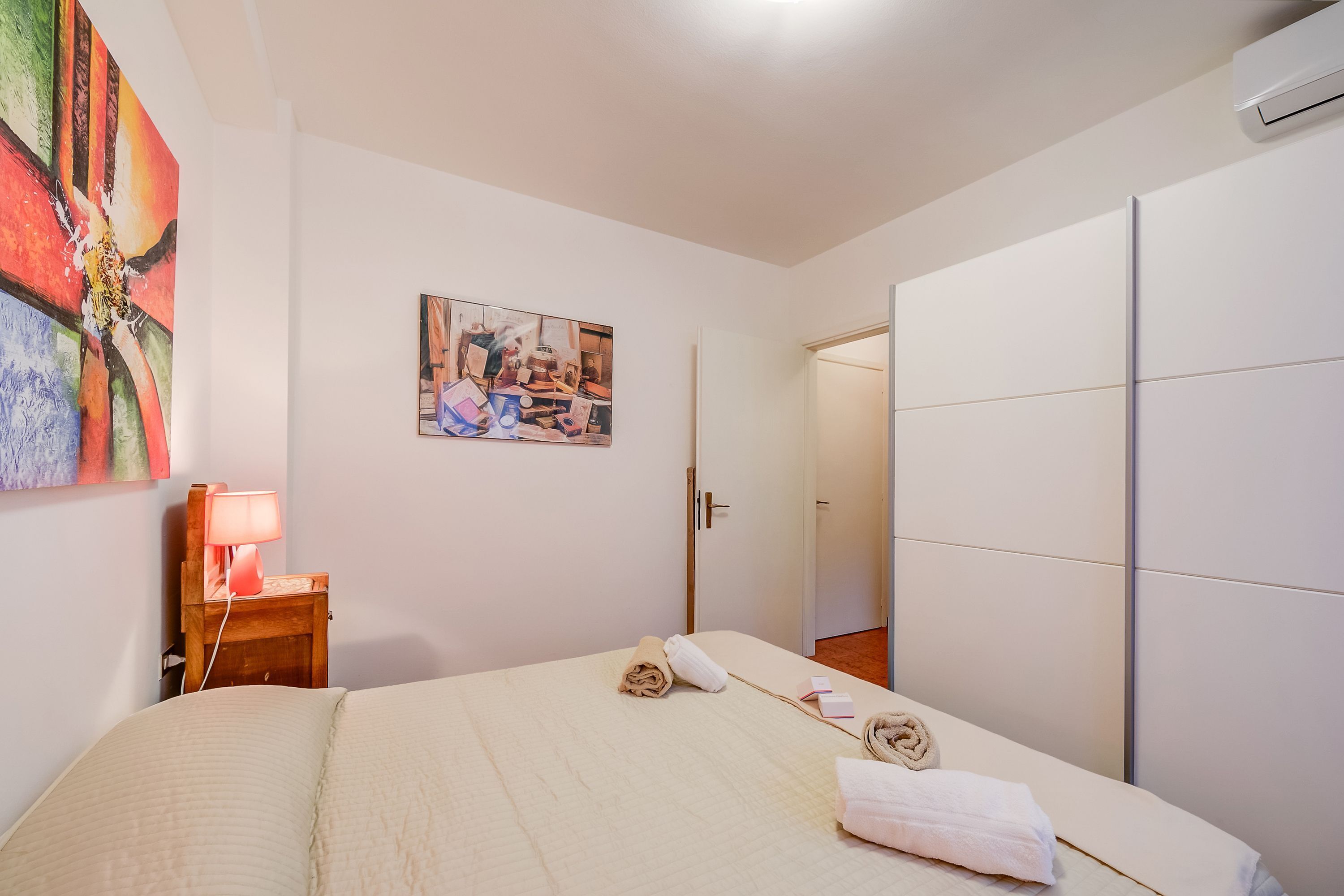 A hotel room with a bed, a nightstand with a lamp, a painting on the wall, and a wardrobe.