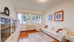 Ein modernes Wohnzimmer mit einem weißen Sofa, einem Glastisch und einem großen Fenster mit Blick auf Bäume.