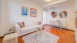 Ein modernes Wohnzimmer mit einem weißen Sofa, einem runden Couchtisch und einem Essbereich mit weißem Tisch und Stühlen.