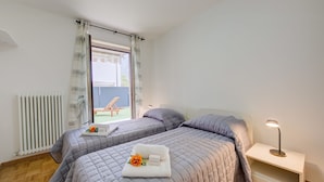 A bedroom with two beds, a bedside table with a lamp, a window with curtains, and a view of the outside. - Casa Karin in Riva del Garda (Riva del Garda)