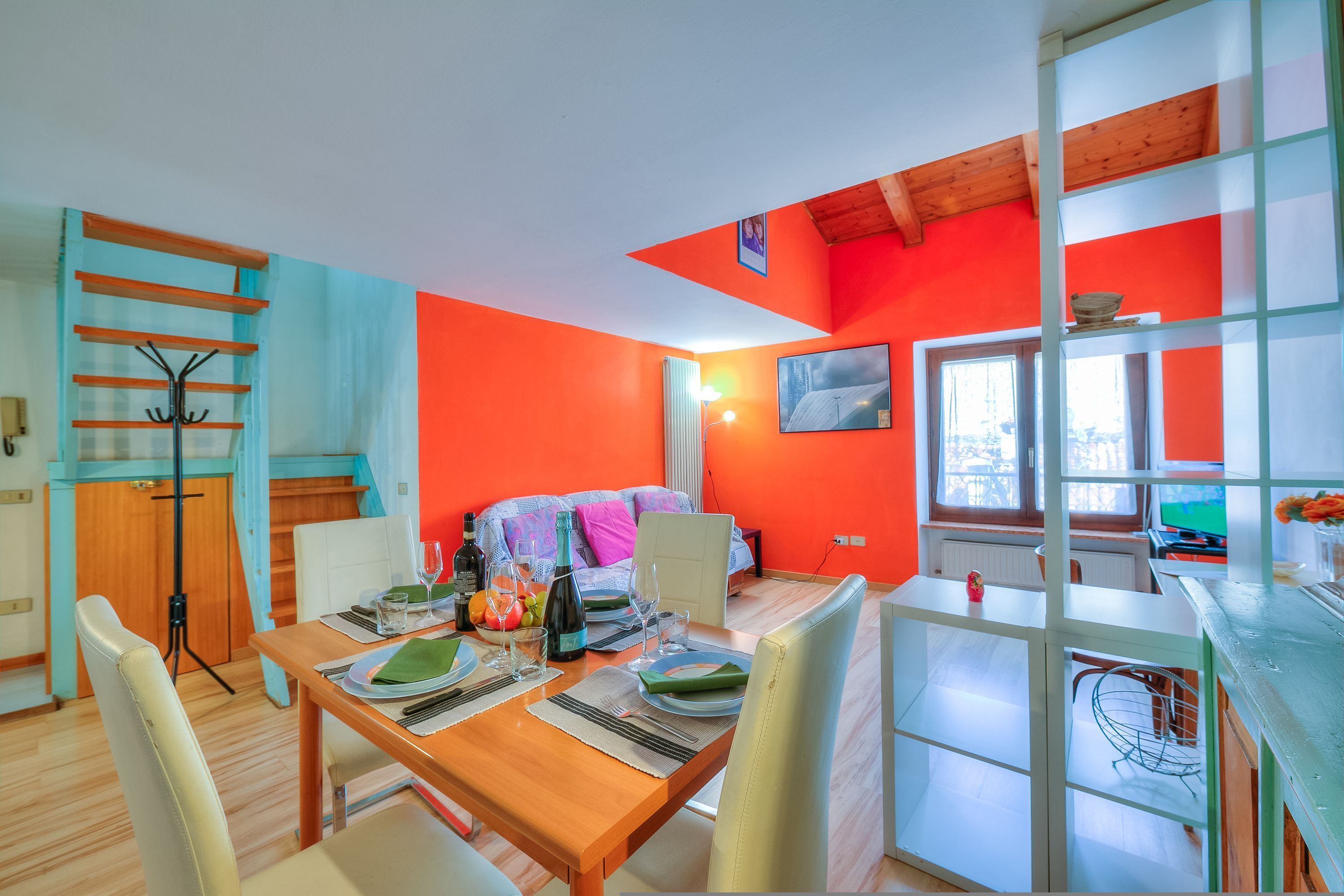 A bright room with orange walls, a wooden dining table, and a bed with a colorful blanket.