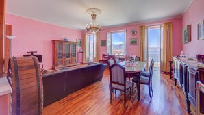 A room with pink walls, wooden flooring, a chandelier, a dining table with chairs, a sofa, and a cabinet. - Il Porticciolo Stresa (Stresa)