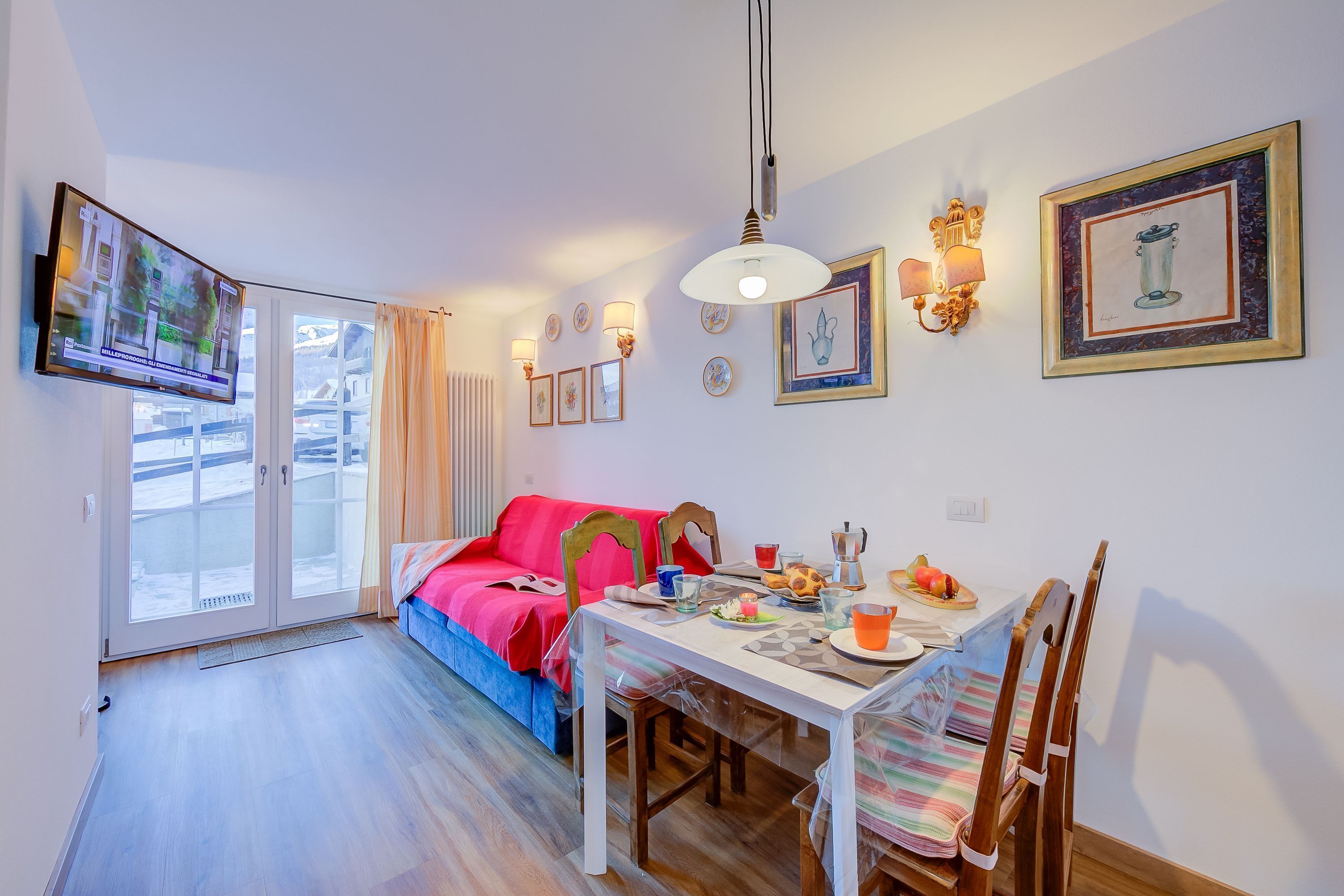 Un coin repas avec une table dressée pour deux personnes, un canapé rouge et un téléviseur fixé au mur.
