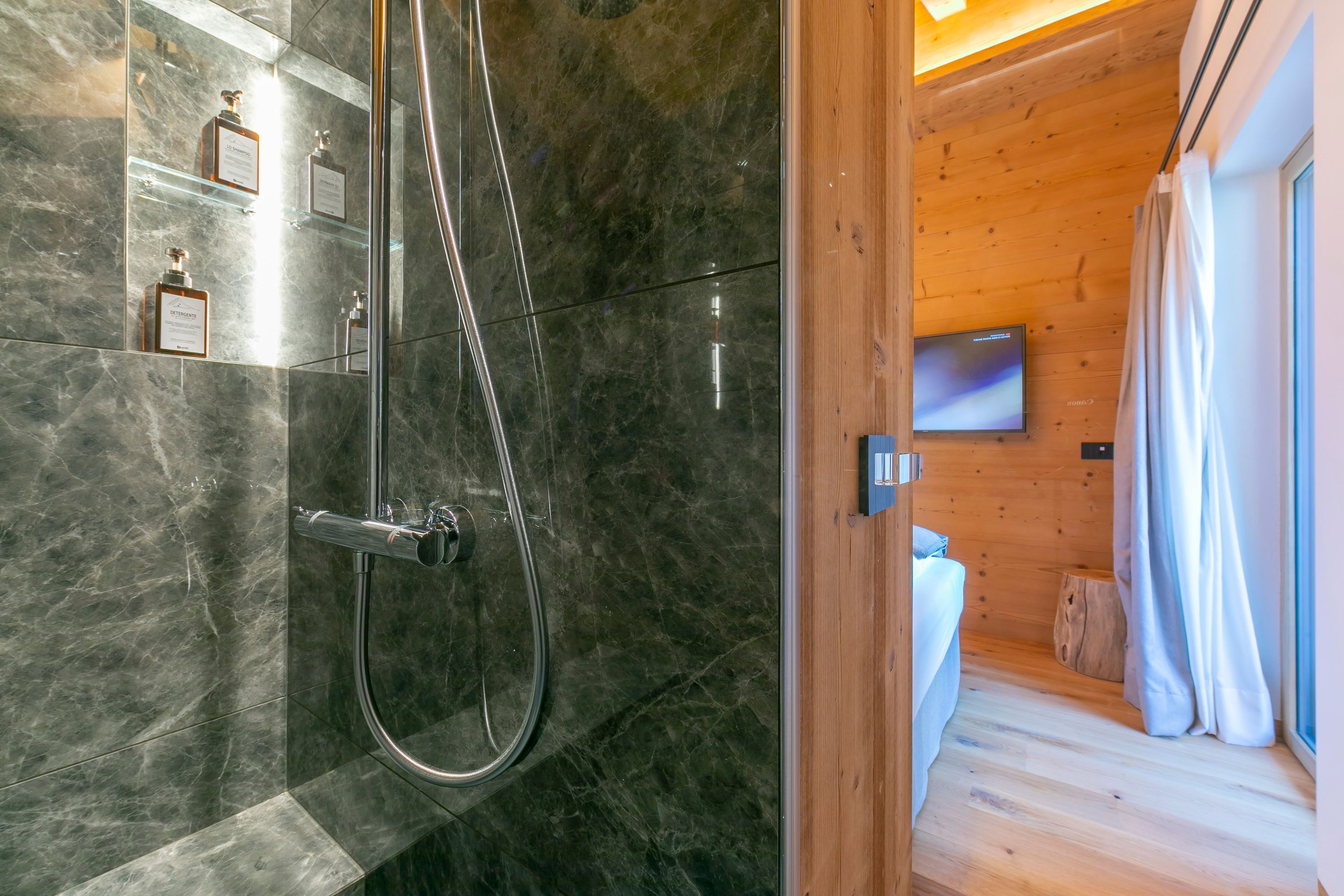A modern bathroom with a glass shower enclosure, marble walls, and a wooden interior visible through an open door.