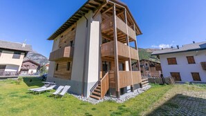 A two-story chalet with a wooden balcony, a stone pathway, and a grassy area with sun loungers. - Giulia in Livigno With 2 Bedrooms and 2 Bathrooms (Livigno)