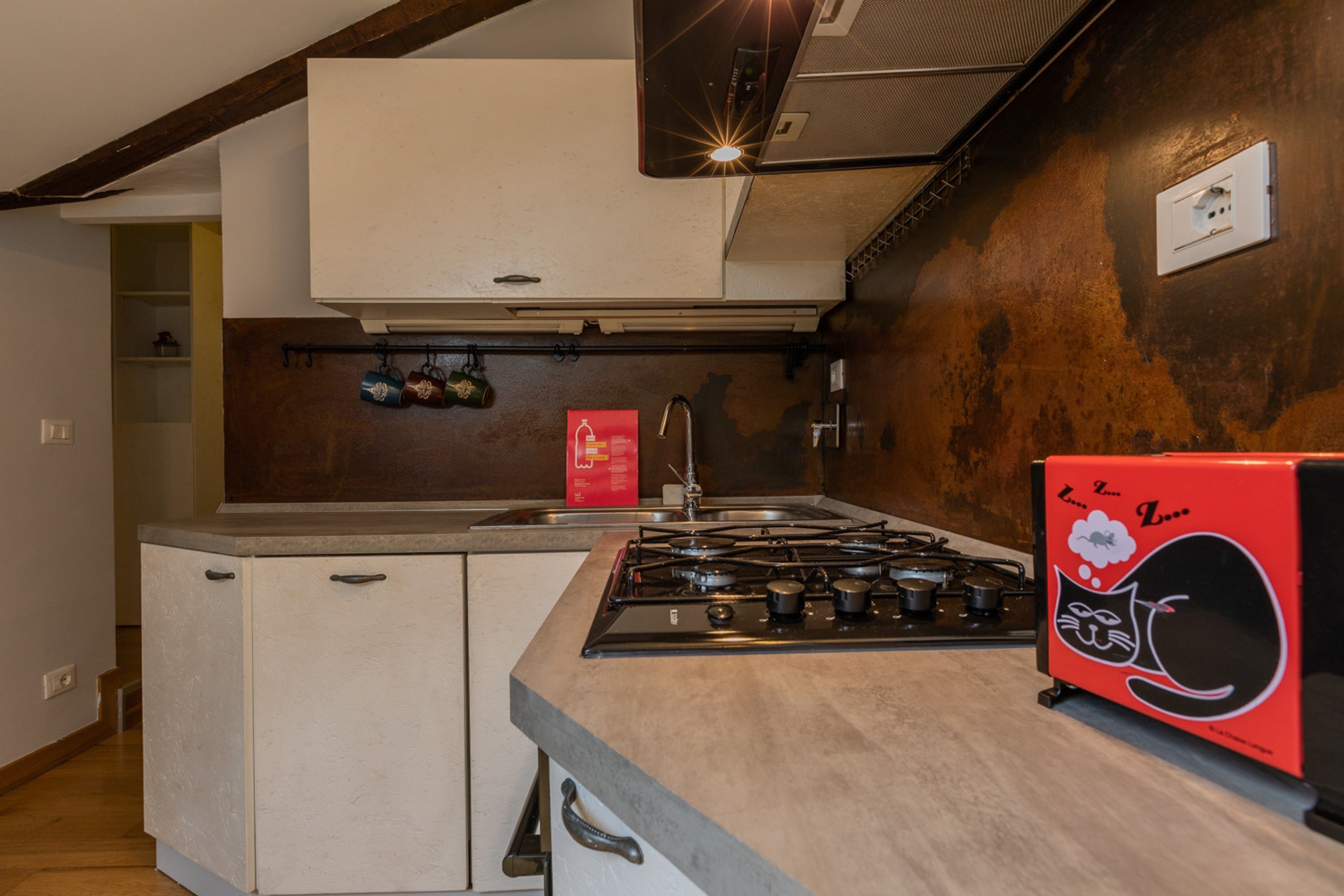 A compact kitchen with a gas stove, white cabinets, and a red appliance with a cat design.
