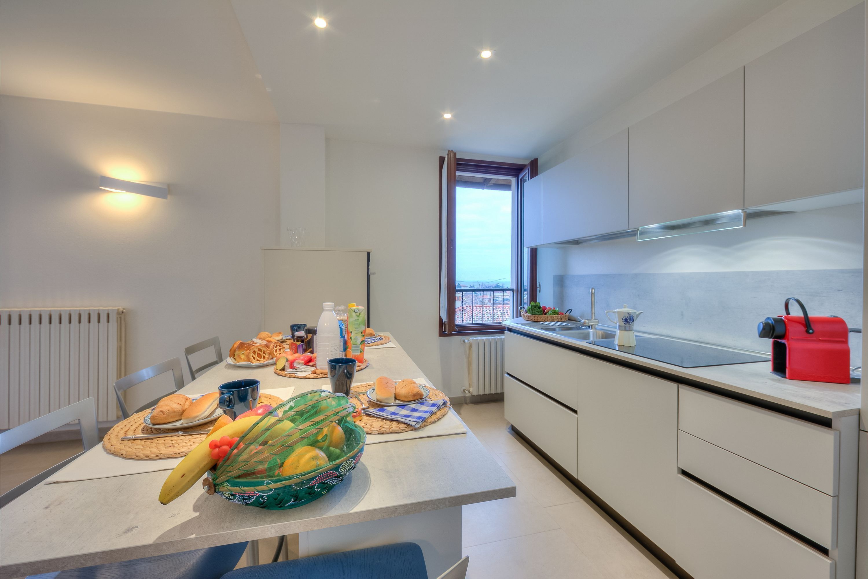 A modern kitchen with a dining table set for breakfast, featuring bread, fruits, and beverages.