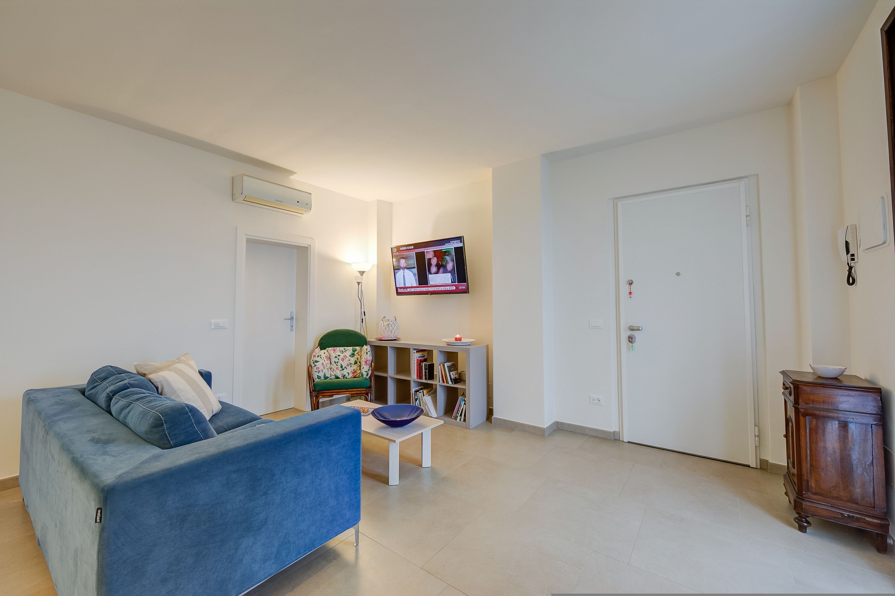 A living room with a blue sofa, a small table, a bookshelf, and a television mounted on the wall.