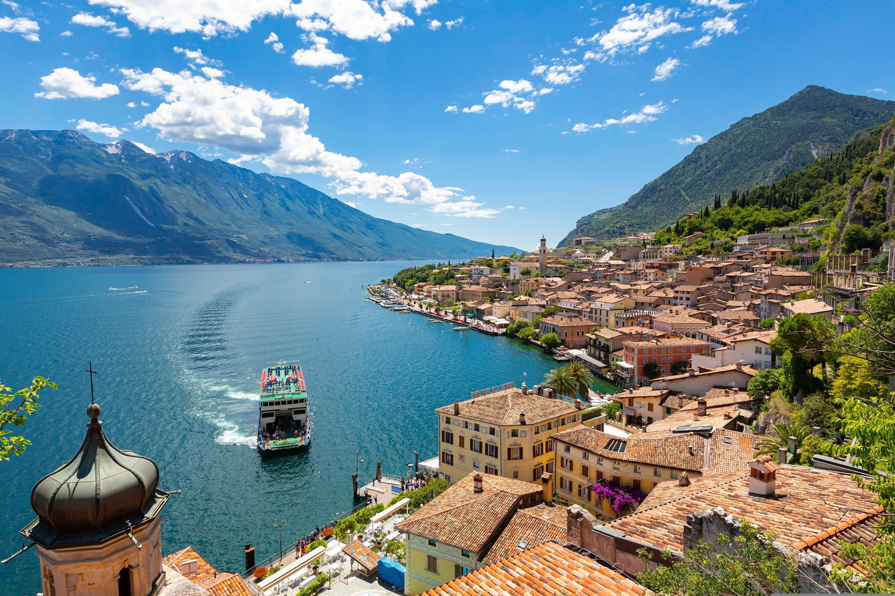 A coastal town with a boat on the water, buildings with red-tiled roofs, and mountains in the background.