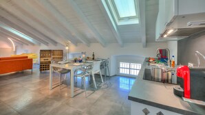 Eine moderne Küche mit Oberlicht, ein Essbereich mit Tisch und Stühlen sowie ein heller, luftiger Wohnraum mit Sofa und Kaffeemaschine.
