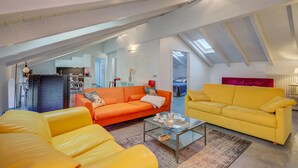 Ein modernes Loft mit hoher Decke, freiliegenden Balken und einem Oberlicht. Es zeigt einen Wohnbereich mit gelben und orangefarbenen Sofas, einem Glastisch und eine Küche im Hintergrund.