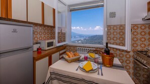 A kitchen with a view of the mountains, a refrigerator, a microwave, a stove, and a table set with bread, wine, and a napkin. - Nicoletta s Lake View On Stresa Hills (Stresa)
