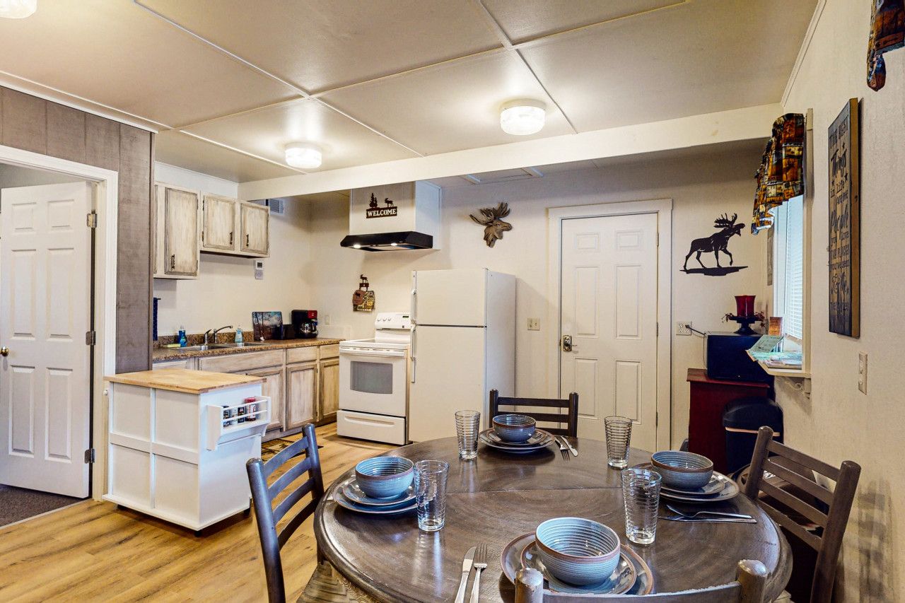 A compact kitchen with white cabinets, a stove, and a refrigerator. There is a dining table set with plates and glasses.