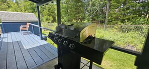 A barbecue grill on a wooden deck with a chair and a small building in the background.