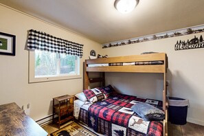 Una cama litera con cubrecolchón a cuadros, una mesita de madera pequeña, un cubo de basura y un letrero de bienvenida en la pared.