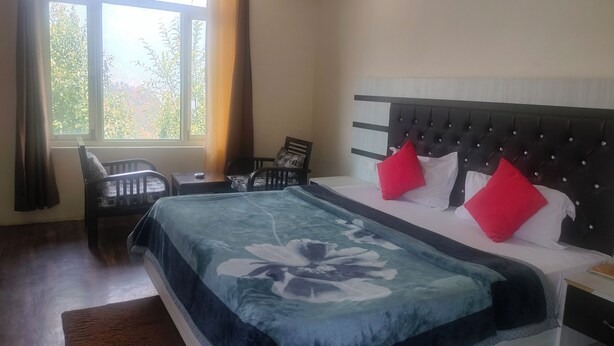 A bedroom with a large bed, a nightstand, a chair, and a window with curtains. - The Shagun Grand (Rohtak)
