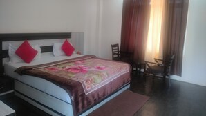 A hotel room with a bed, a nightstand, a chair, a small table, and a window with curtains. - The Shagun Grand (Rohtak)