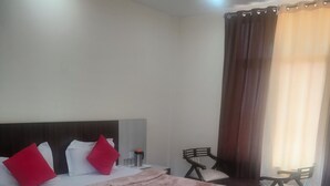 A neatly made bed with a patterned bedspread, a bedside table with a lamp, a chair, and a window with curtains. - The Shagun Grand (Rohtak)