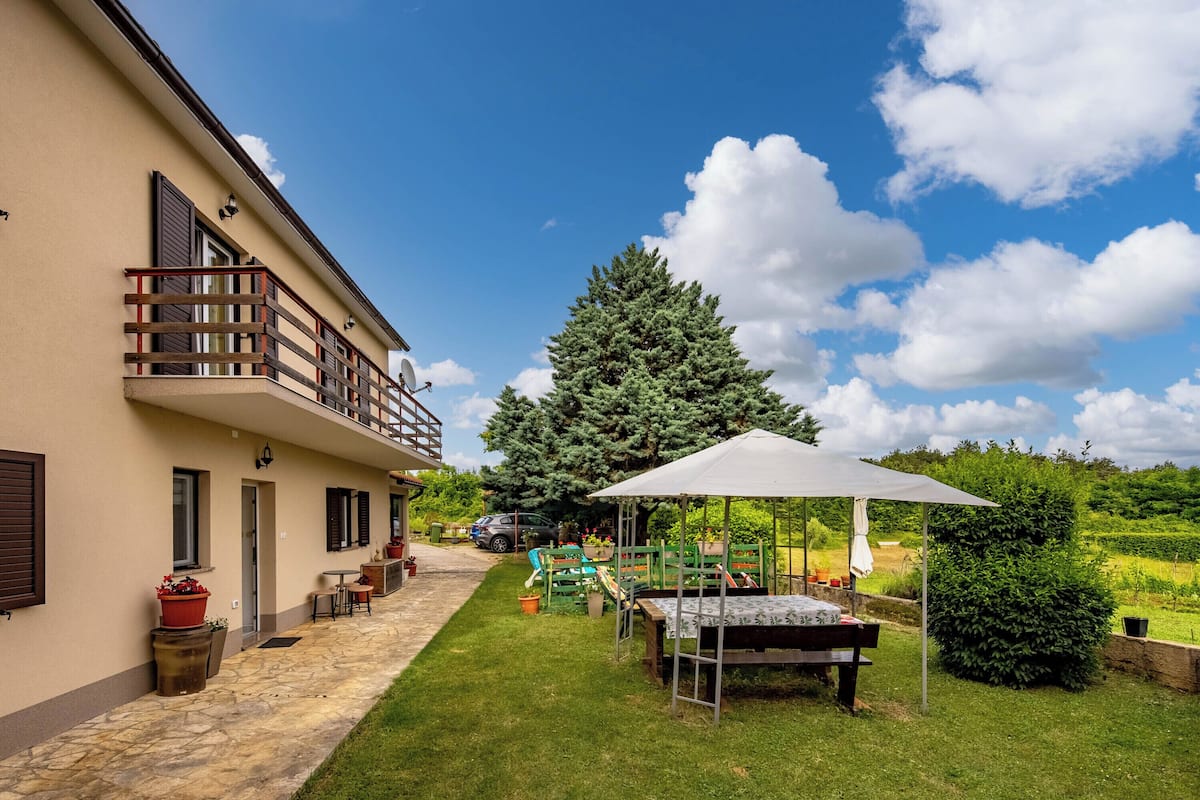 A house with a balcony, a patio with a table and chairs, and a garden area with a large tree and a gazebo.