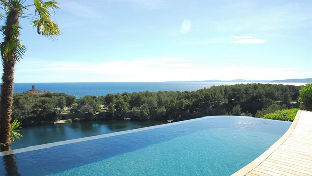 A clear blue infinity pool with a wooden deck, surrounded by lush greenery and overlooking a body of water with distant land.