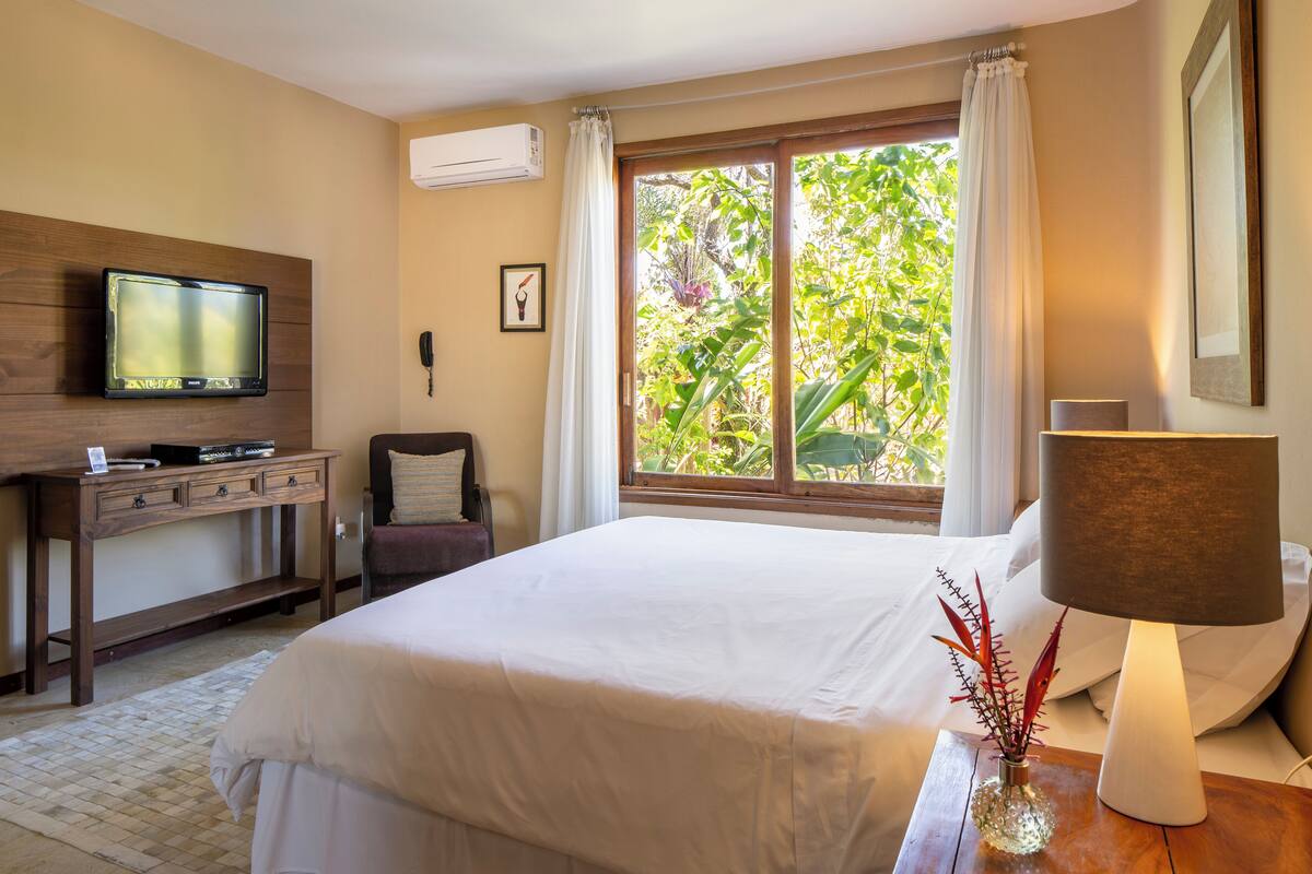Una habitació d'hotel amb un llit, una taula de treball amb un ordinador, un televisor, una finestra amb cortines i una làmpada.
