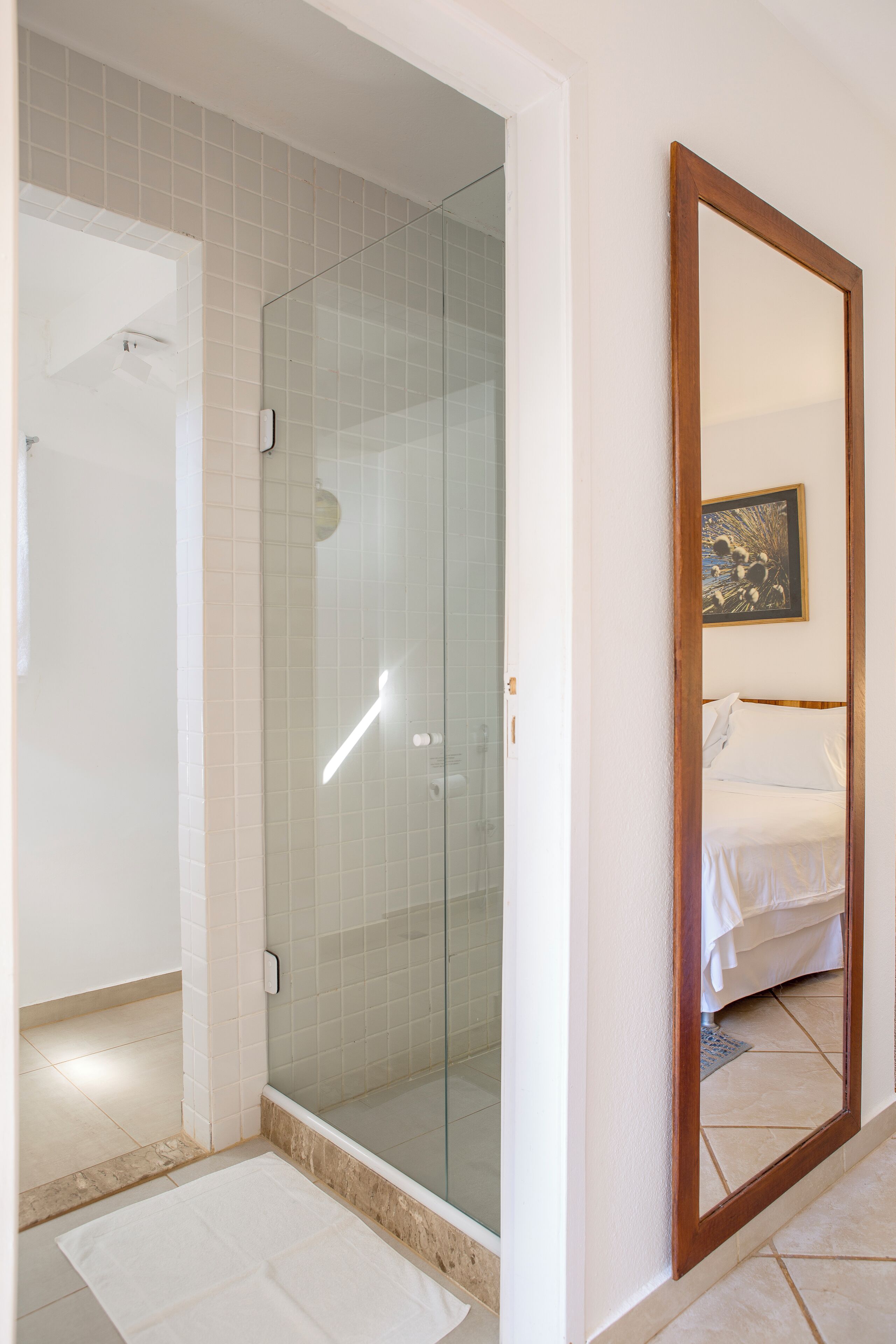 A bathroom with a glass-enclosed shower, a white sink, and a large mirror reflecting a bed with white bedding.