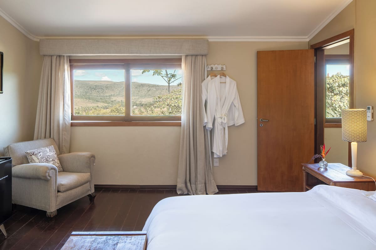 Una habitació d'hotel amb llit, cadira, escriptori, finestra amb cortines, una bata penjada a la paret i una làmpada de nit.