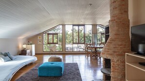 A spacious loft bedroom with a curved bed, a blue ottoman, a stone fireplace, and a TV on a wooden shelf. - Pousada Maya (Alto Paraíso de Goiás)