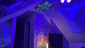 Una habitación con techo inclinado, un televisor de pantalla plana sobre un mueble bajo y una pared con un mural abstracto y colorido.
