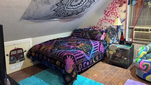 Un dormitorio con una cama, un tapiz colorido, una lámpara y una mesita con objetos decorativos.