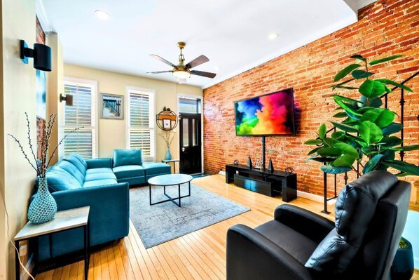 A living room with a brick wall, a ceiling fan, a TV, a sofa, a coffee table, and a plant.