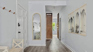 Un pasillo con piso de madera, paredes blancas y una puerta con espejo. Hay obras de arte enmarcadas en la pared y un marco de madera alrededor de la puerta.