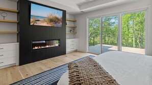 A modern bedroom with a fireplace, a large TV, and a balcony with a view of trees. - Juniper Presents: Contemporary 26-Acre Estate, Private Beach, Pool (Stevensville)