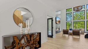 A modern living room with a large window, a round mirror, and a console table with a wooden sculpture. - Juniper Presents: Contemporary 26-Acre Estate, Private Beach, Pool (Stevensville)