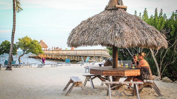 Ein Strand mit einer strohgedeckten Struktur, Holzbänken und einem Holztisch.