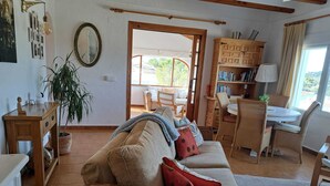 A cozy living room with a sofa, a wooden cabinet, a dining table with chairs, and a large window. - Villa Bibi with Spectacular Sunset and Views (Javea)