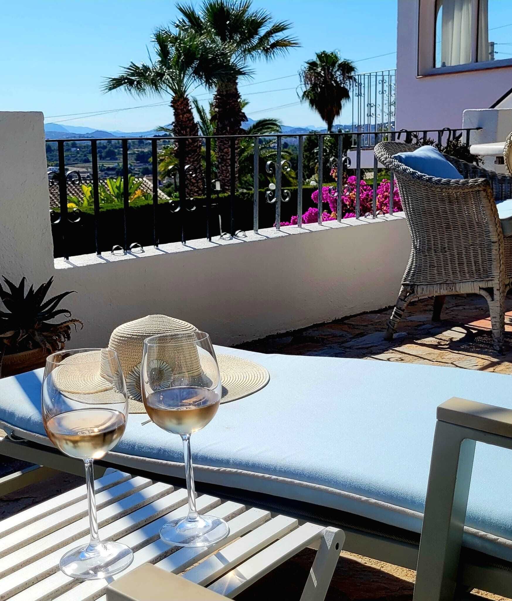 A balcony with a white cushioned lounge chair, a wicker chair, and two wine glasses on a table.