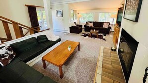 A spacious living room with a large sofa, a coffee table, and a fireplace. There is a staircase leading to another level, and a TV mounted on the wall.