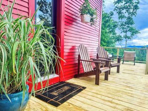 Een houten terras met een rood gebouw, twee Adirondack stoelen, een welkomstmat en een hangplant met witte bloemen.