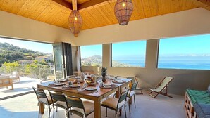 A dining area with a table set for four, surrounded by chairs, with a view of the sea and hills in the background. - Villa San Joseph (Cargès)