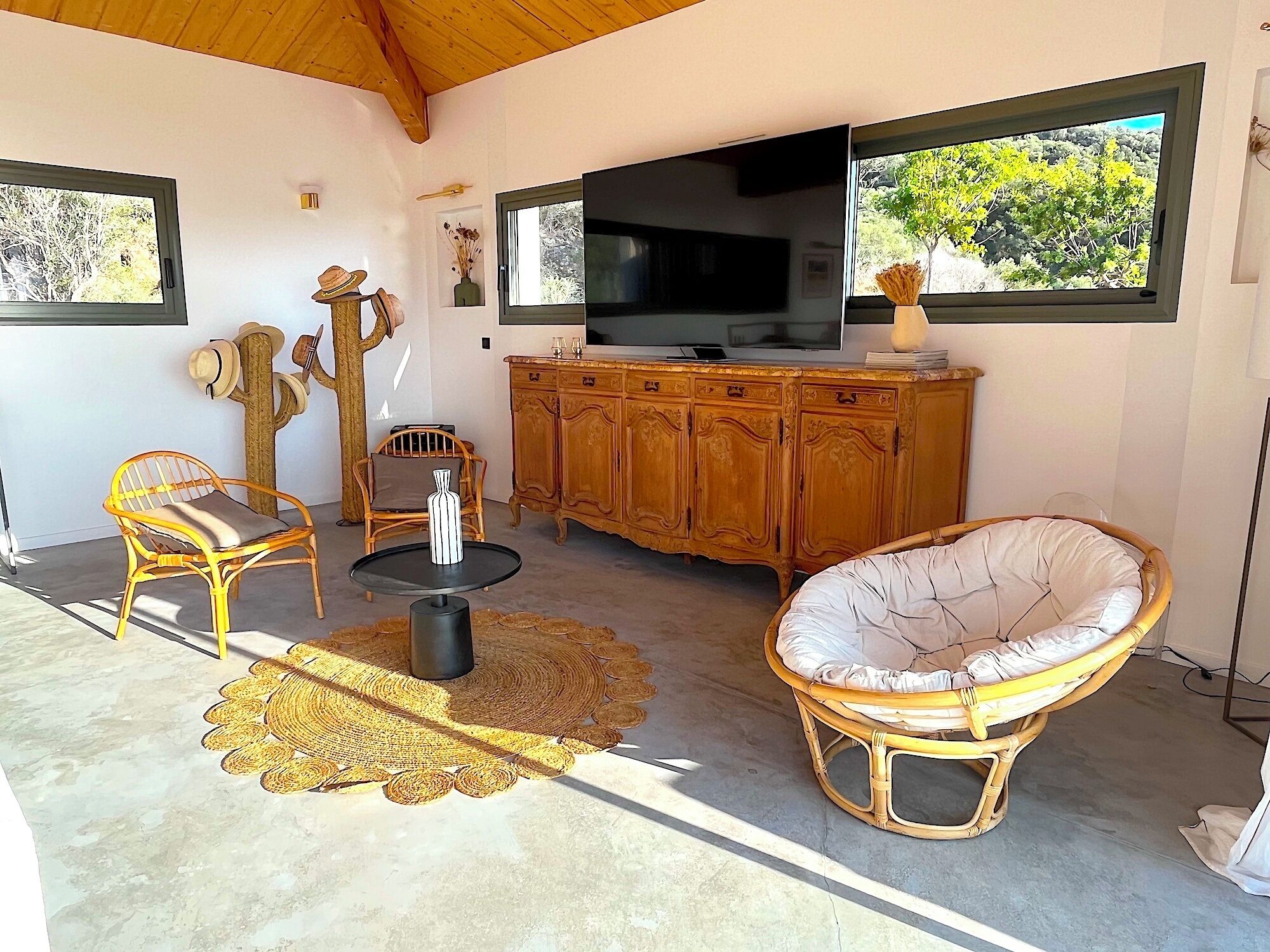 Un salon avec un téléviseur, des meubles en osier, un vase décoratif et une vue sur la verdure par les fenêtres.