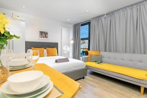 Een moderne hotelkamer met een bed, een bank, een eettafel met borden en glazen, en een raam met gordijnen.