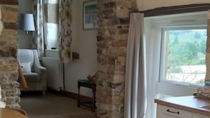 A spacious room with a wooden floor, stone walls, a dining area with a table and chairs, a kitchen with white cabinets, and a view of greenery outside.