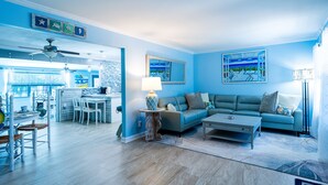 Un salon avec un canapé bleu, une table à manger avec des chaises, et un coin cuisine à l’arrière-plan.