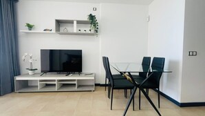 A modern living room with a flat-screen TV, a glass dining table with four chairs, and a white entertainment unit with shelves. - Casa Rigola El Cotillo (El Cotillo)