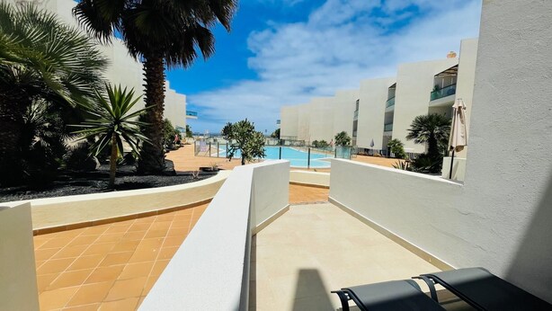 A pool area with tiled flooring, palm trees, and a clear blue sky. - Casa Rigola El Cotillo (El Cotillo)