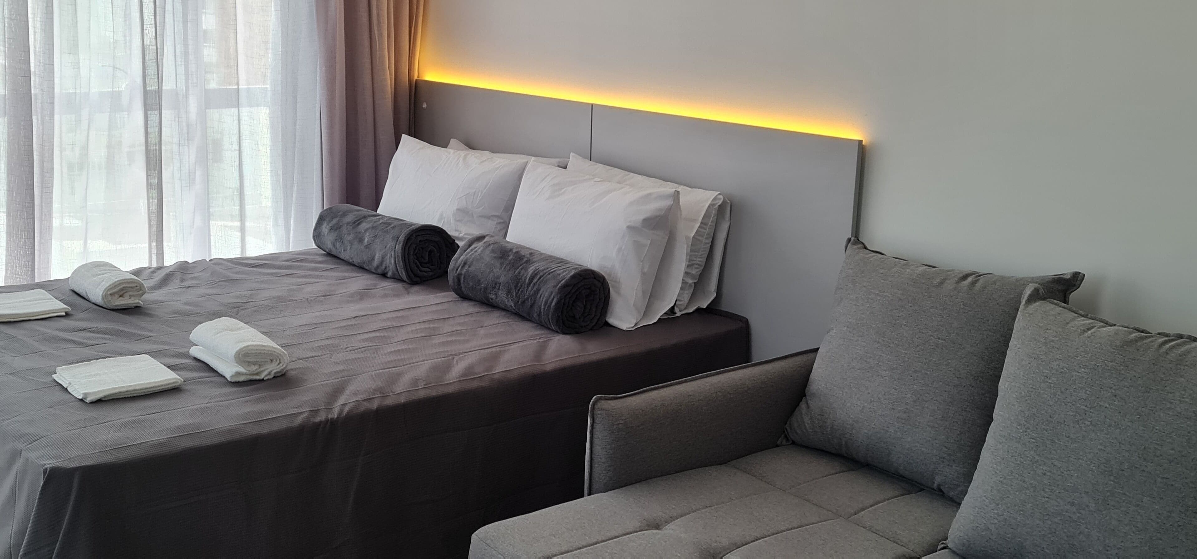 Habitación de hotel con una cama bien hecha, un sofá y un cabecero con iluminación integrada.