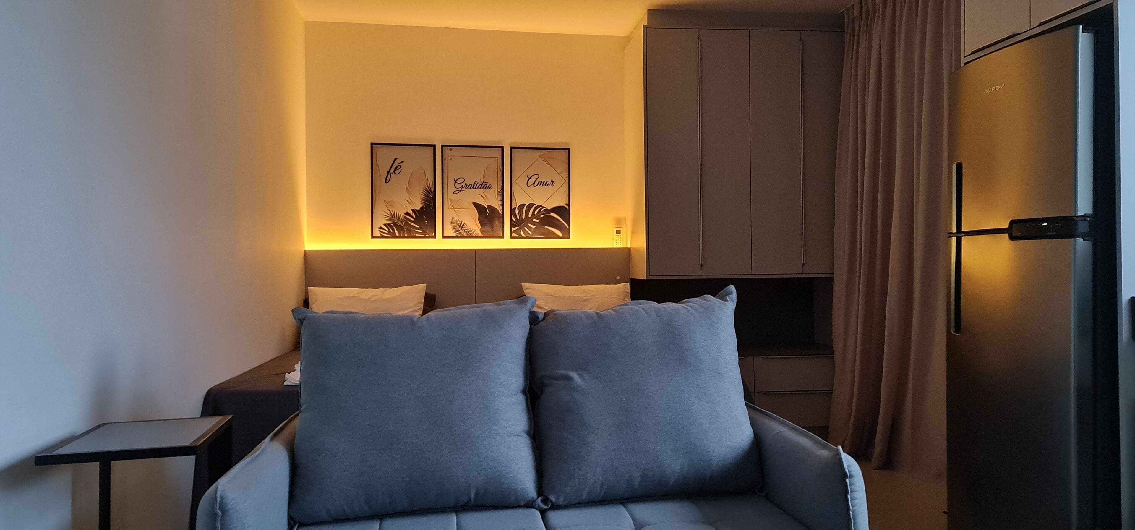 Una habitación de hotel moderna con un sofá gris, una mesita auxiliar pequeña y un mueble-vitrina de pared. Encima del sofá hay tres obras de arte enmarcadas.